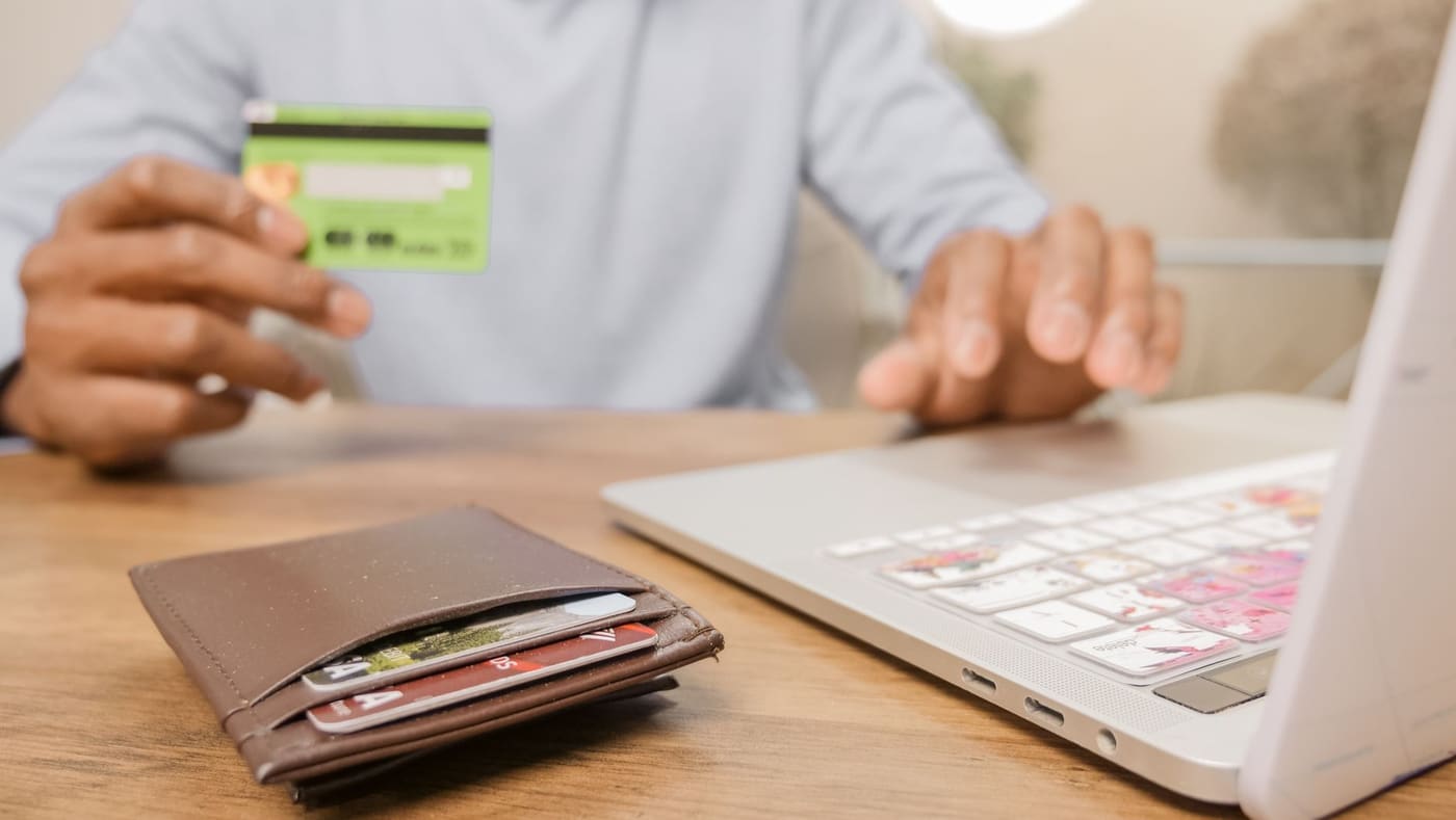 A hand holding a bank card beside a laptop and a wallet