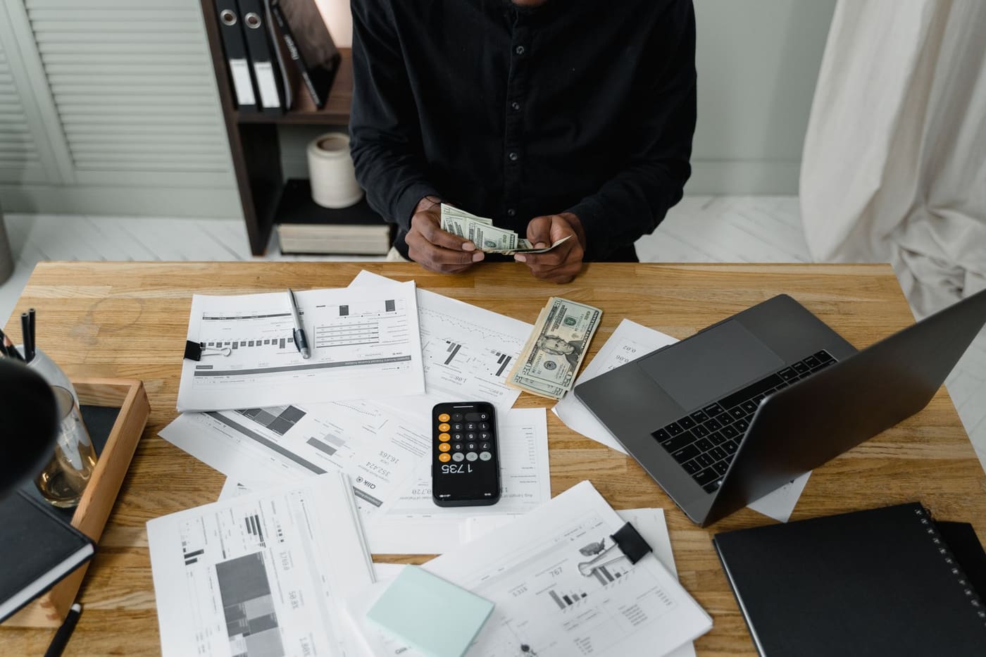 A person counting dollar bills at a desk with a laptop, papers and calculator