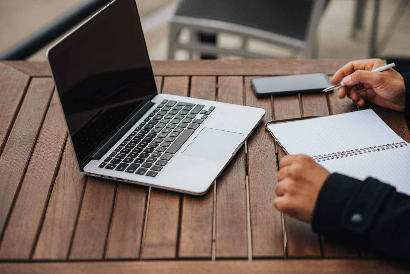 A laptop, a smartphone and a spiral notebook on a wooden outdoor table