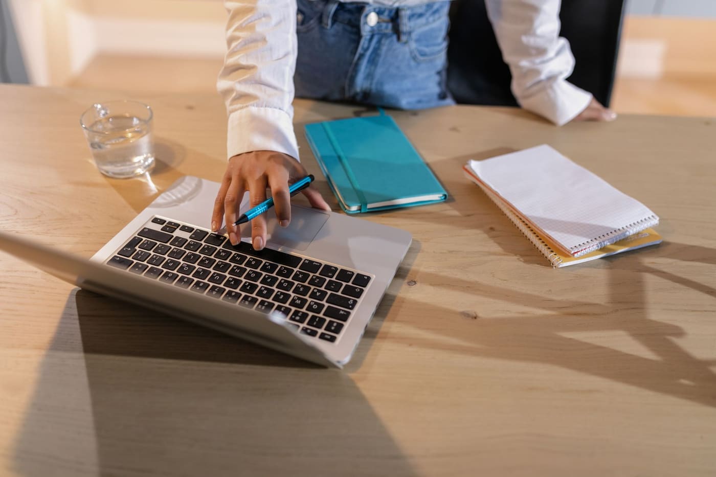 Overhead view of a person using a laptop on a wooden desk with notebooks and a glass of water